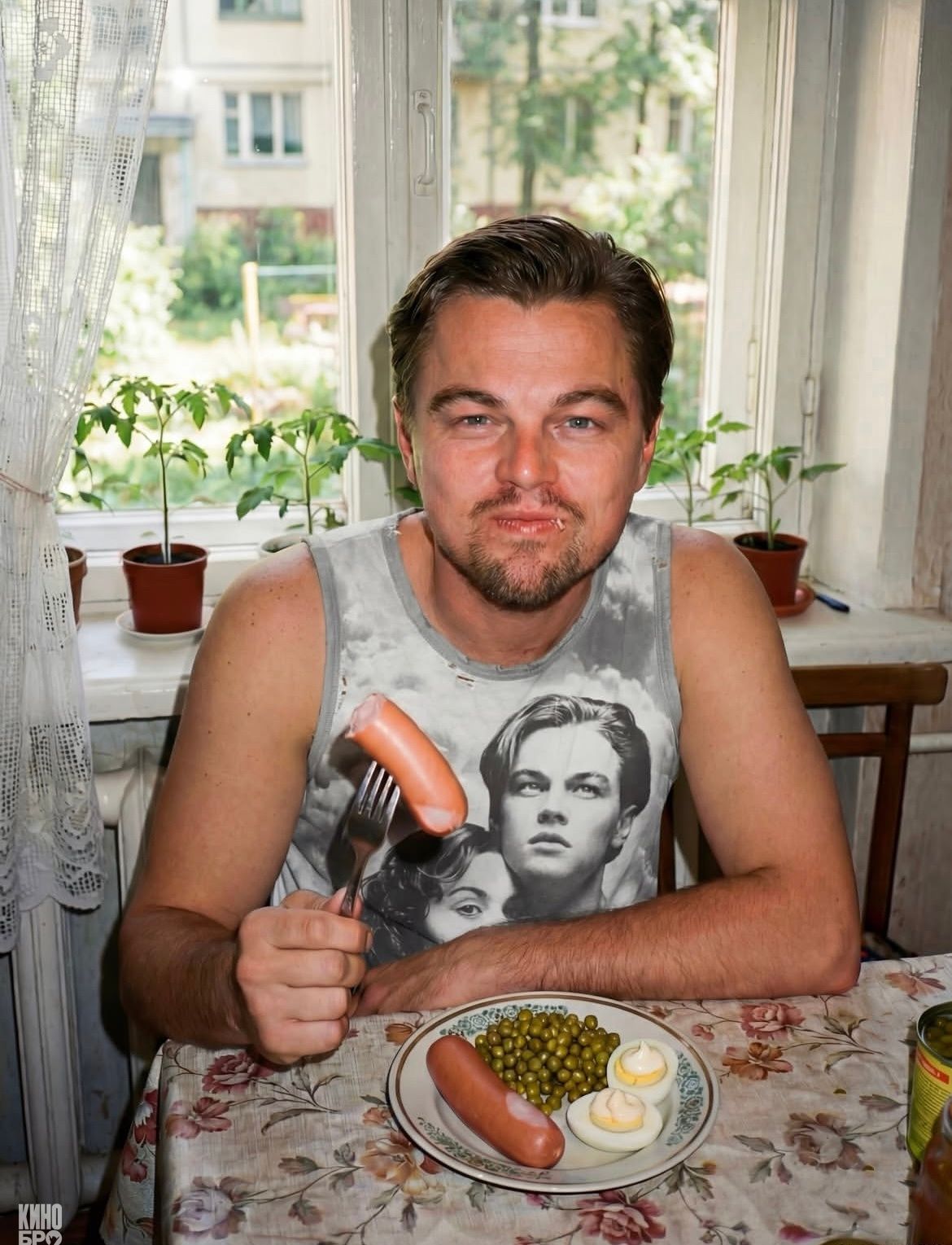 Мужчина в серой майке держит сосиску на вилке за столом. На тарелке горошек и половинки варёного яйца.