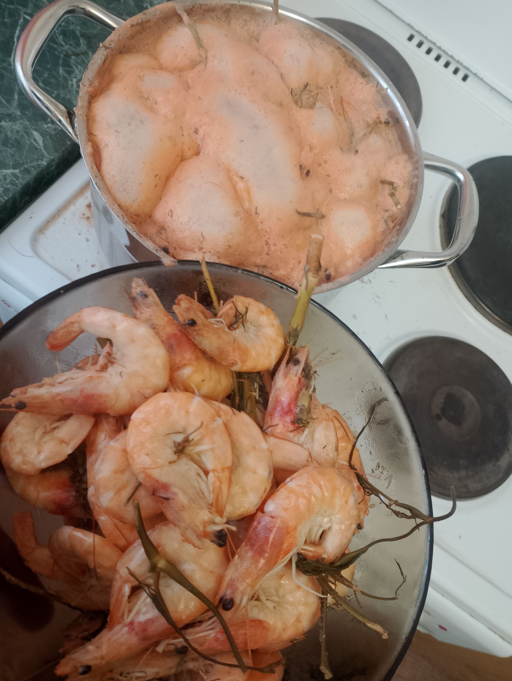 На кухне две кастрюли: в одной варятся креветки, в другой лежат очищенные креветки с хвостами и травами.