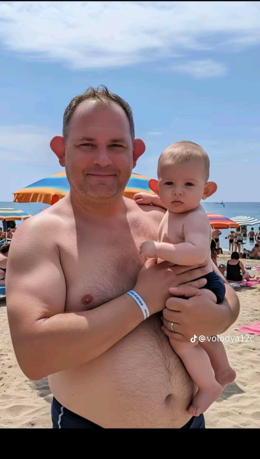 На пляже мужчина держит на руках ребенка. На мужчине нет футболки, на ребенке плавки. На заднем плане видны зонтики, люди, море и небо.