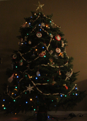 A Christmas tree decorated with garlands, multicolored lights, ornaments, and a star topper. The room is dimly lit.