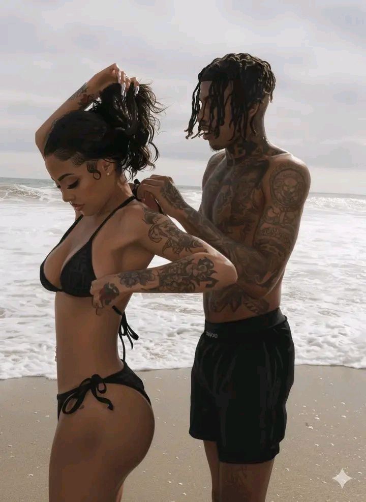 A tattooed man stands behind a woman on the beach, adjusting her black bikini top as she ties it.