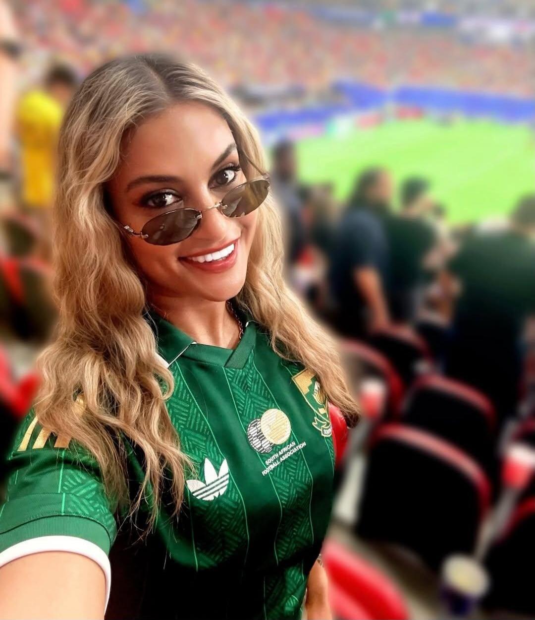A woman with blonde hair and sunglasses is smiling at the camera, wearing a green football jersey with 'SOUTH AFRICAN FOOTBALL ASSOCIATION' and an Adidas logo visible. She is in a stadium with a blurred background showing a sports field and other spectators.