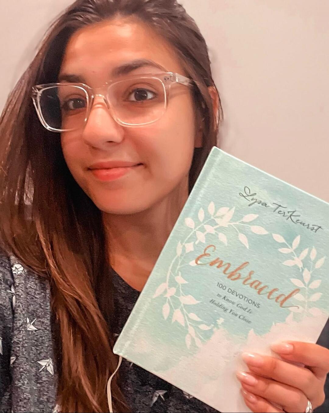 A woman with glasses is smiling and holding a book titled 'Embraced' by Lysa TerKeurst, which says '100 DEVOTIONS to Know God Is Holding You Close'.