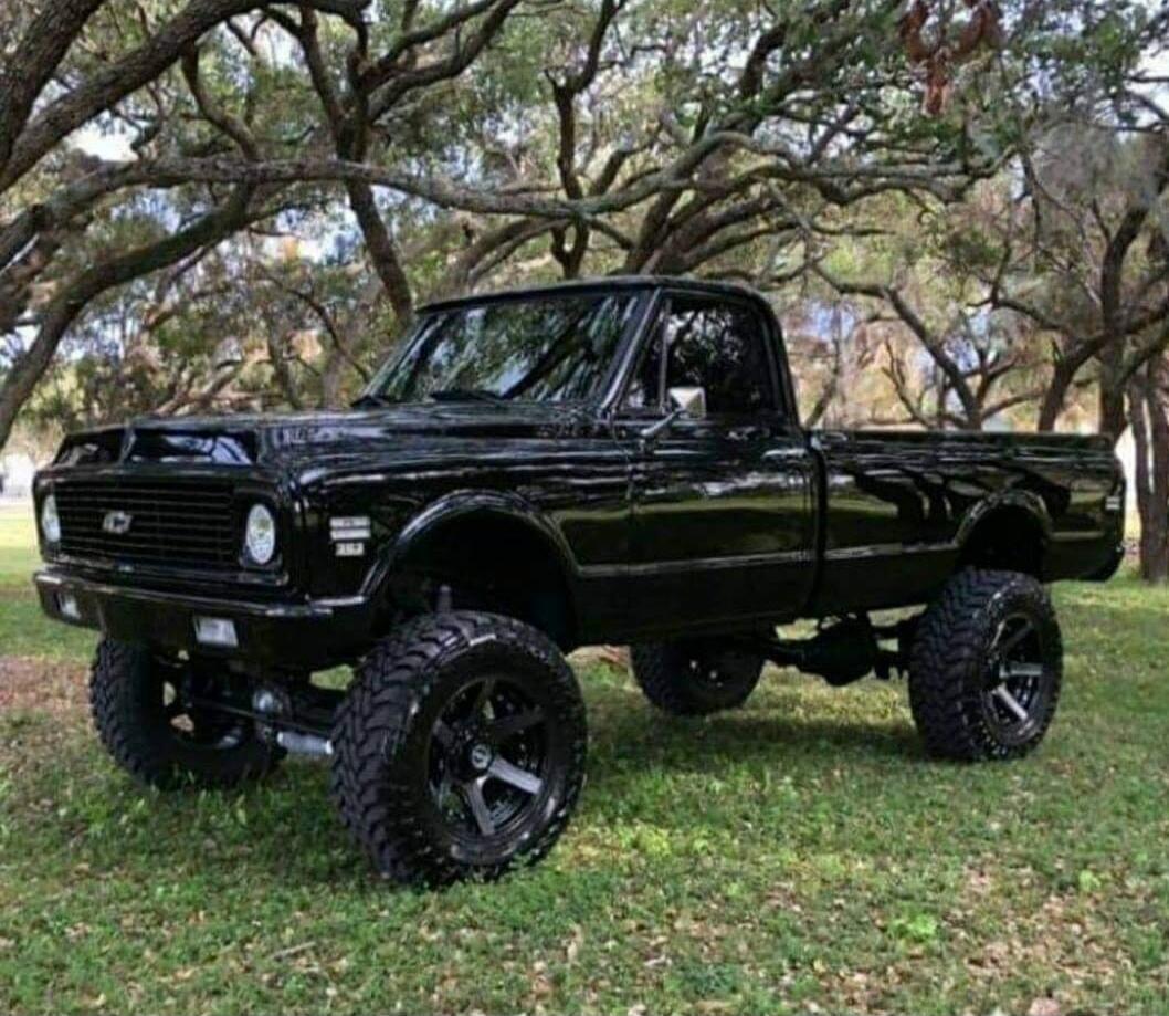 A lifted black vintage pickup truck with oversized off-road tires parked on grass under trees.