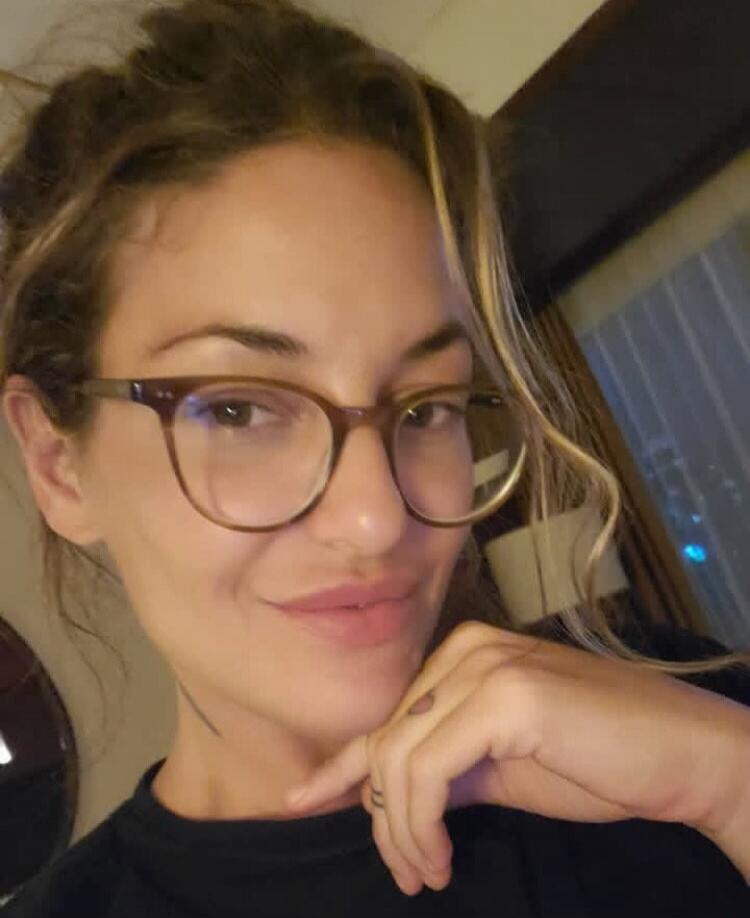 A woman wearing glasses and smiling, resting her chin on her hand.