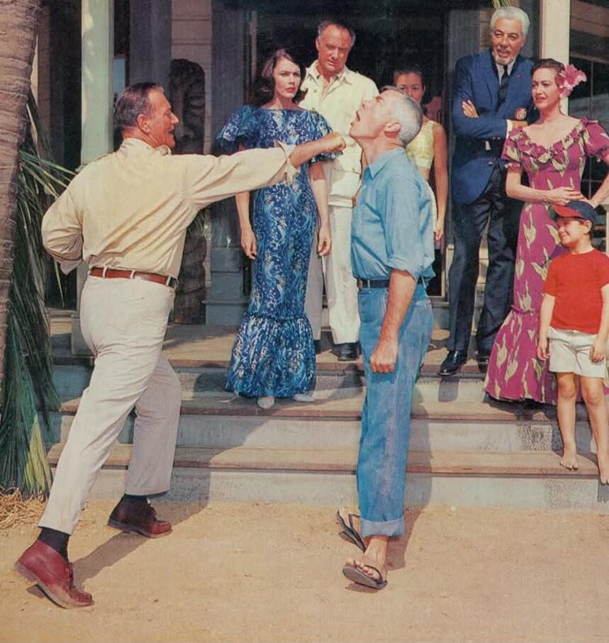 A man in a light shirt and white pants appears to be punching another man in a blue shirt and jeans. Several other people are watching from the steps of a building in the background. The scene looks theatrical or staged, possibly from a movie or play.