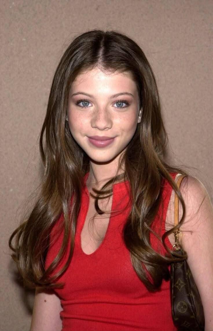 A young woman with long brown hair wearing a red sleeveless top, standing against a beige wall.