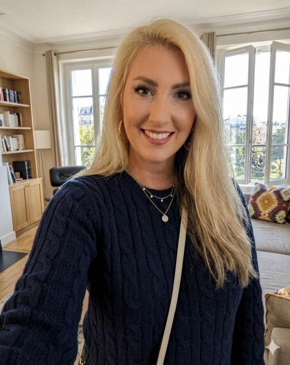 A smiling blonde woman in a dark blue cable-knit sweater takes a selfie indoors. She has long blonde hair and is wearing multiple layered necklaces and hoop earrings. In the background, there's a bright room with a bookshelf, a large window overlooking an urban scene, and a sofa with patterned cushions.