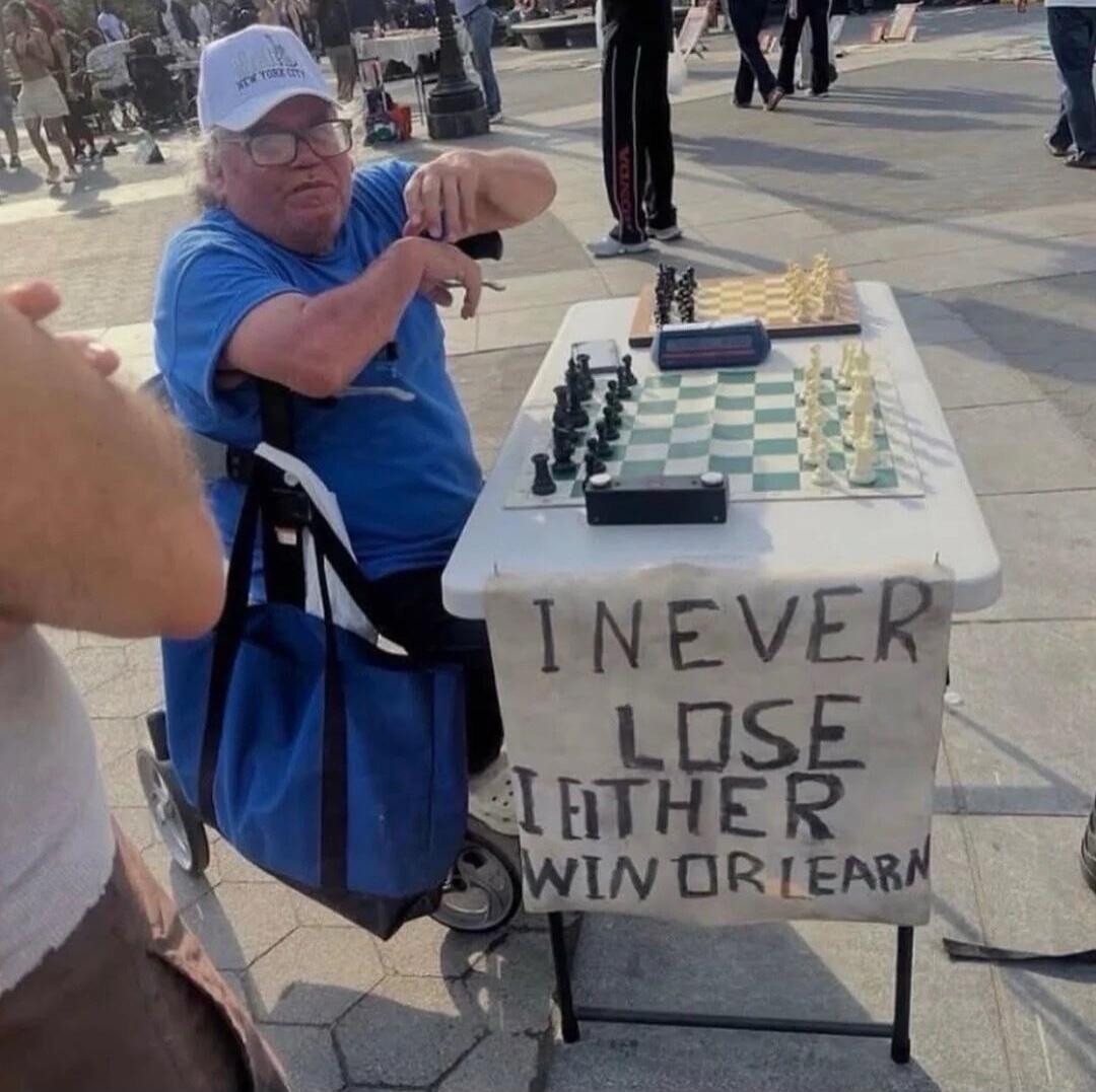 I NEVER LOSE I EITHER WIN OR LEARN. A man in a wheelchair playing chess in a public place, wearing a cap that says 'NEW YORK CITY'.