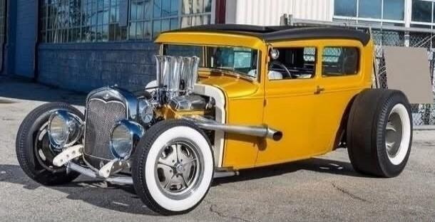 A custom-built yellow hot rod car with large chrome engine components and wide white-wall tires is parked outdoors. The car has a classic design with a modern custom look.