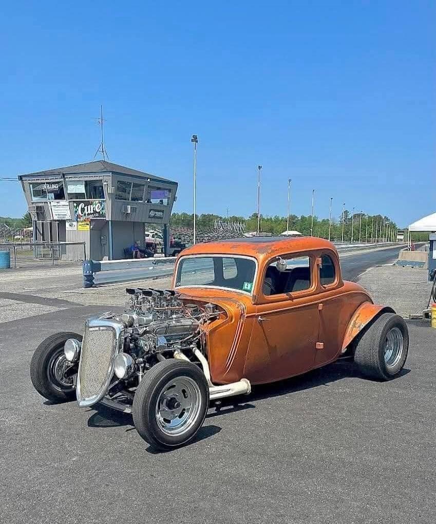 Orange vintage hot rod with a large exposed engine parked on a race track. A two-door coupe with a classic retro look, chrome components, and big rear tires. Background shows a small building/booth and track facilities.