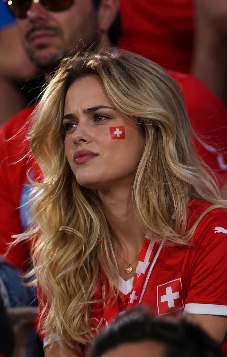 A young woman with blonde, wavy hair, wearing a red sports jersey with a Swiss flag emblem. She has a small Swiss flag painted on her cheek and is looking intently, possibly watching a game at a stadium.