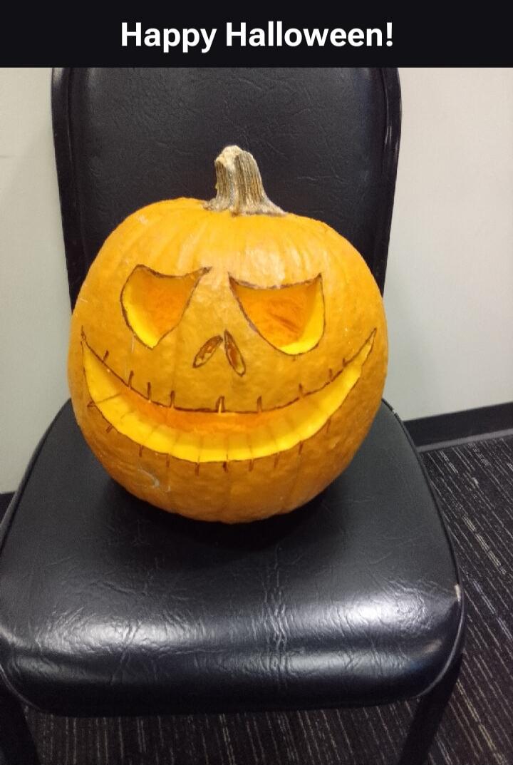 Happy Halloween! A carved pumpkin, also known as a jack-o'-lantern, with a spooky face is sitting on a black office chair.