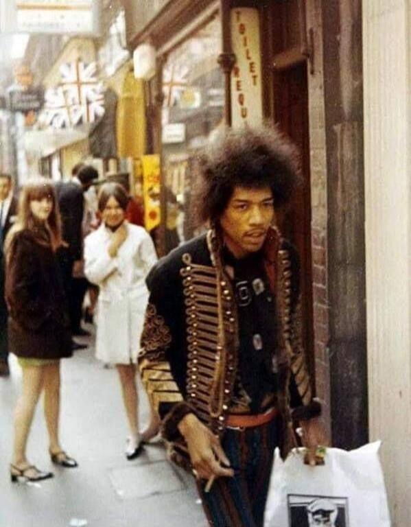 A vintage photograph of Jimi Hendrix walking down a street, holding a shopping bag and a cigarette. He is wearing a distinctive military-style jacket and striped trousers. Other people and Union Jack flags are visible in the background. A sign partially reads 'TOILET REGUL'.