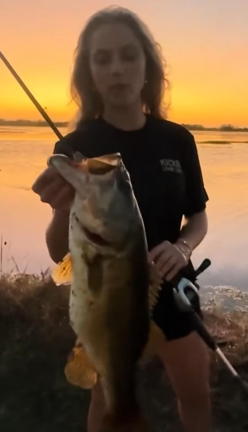 A person standing by a body of water at sunset, holding up a large fish with a fishing rod in the background.