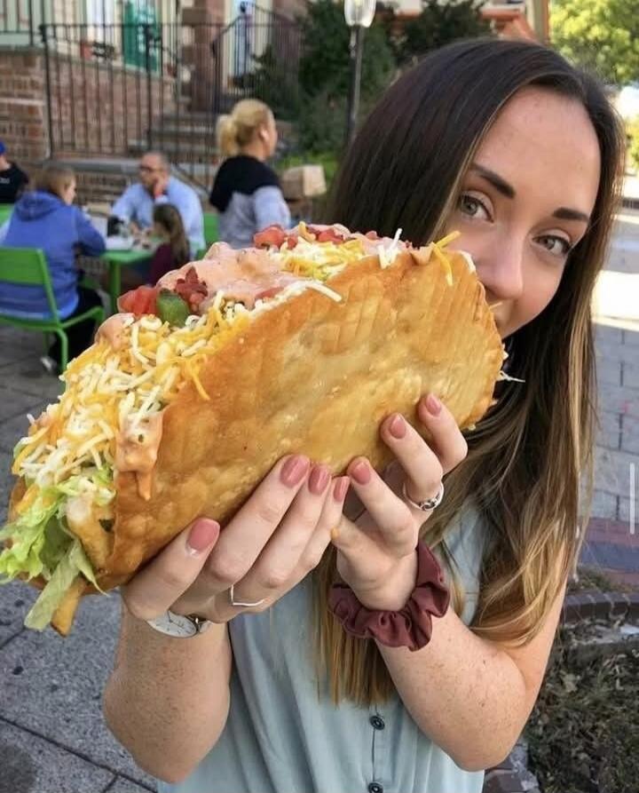 A woman is holding a massive taco/burrito filled with lettuce, cheese, and sauce outside, smiling at the camera.
