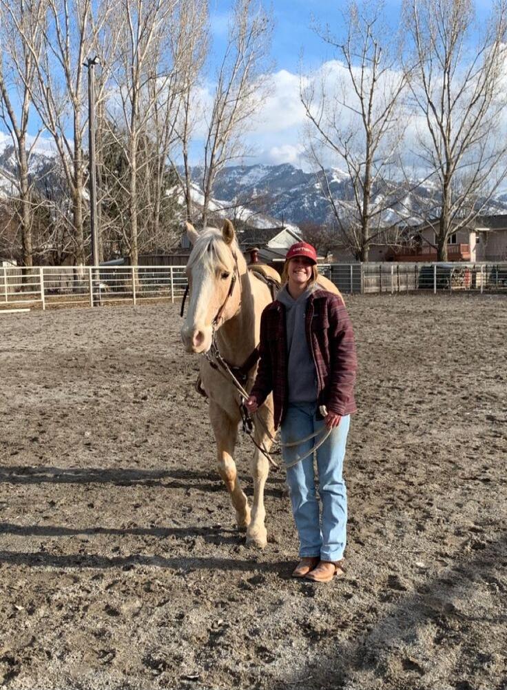 A person standing next to a beige horse in an outdoor arena, with mountains and bare trees in the background.
