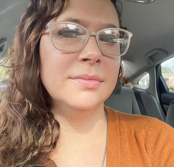 A woman wearing glasses sits in a car, with wavy hair and an orange top.