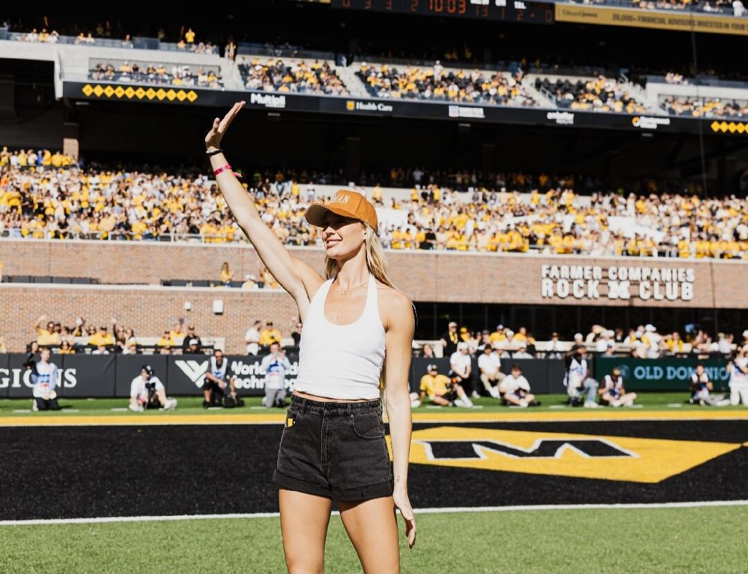 A woman on a sports field, possibly a football stadium, waving to a large crowd of spectators. Signs visible include 'FARMER COMPANIES ROCK 'N' CLUB', 'OLD DOMINION', and 'Health Care'. A scoreboard shows '10:03' and scores '13 7 0'.