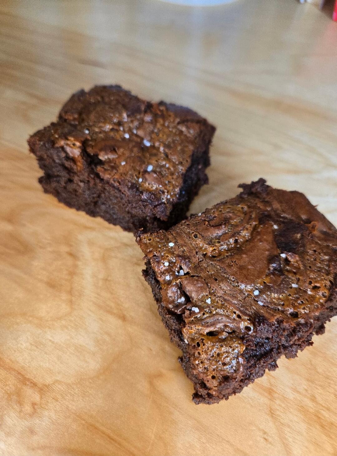 Two pieces of chocolate brownies with sea salt flakes on a wooden surface.