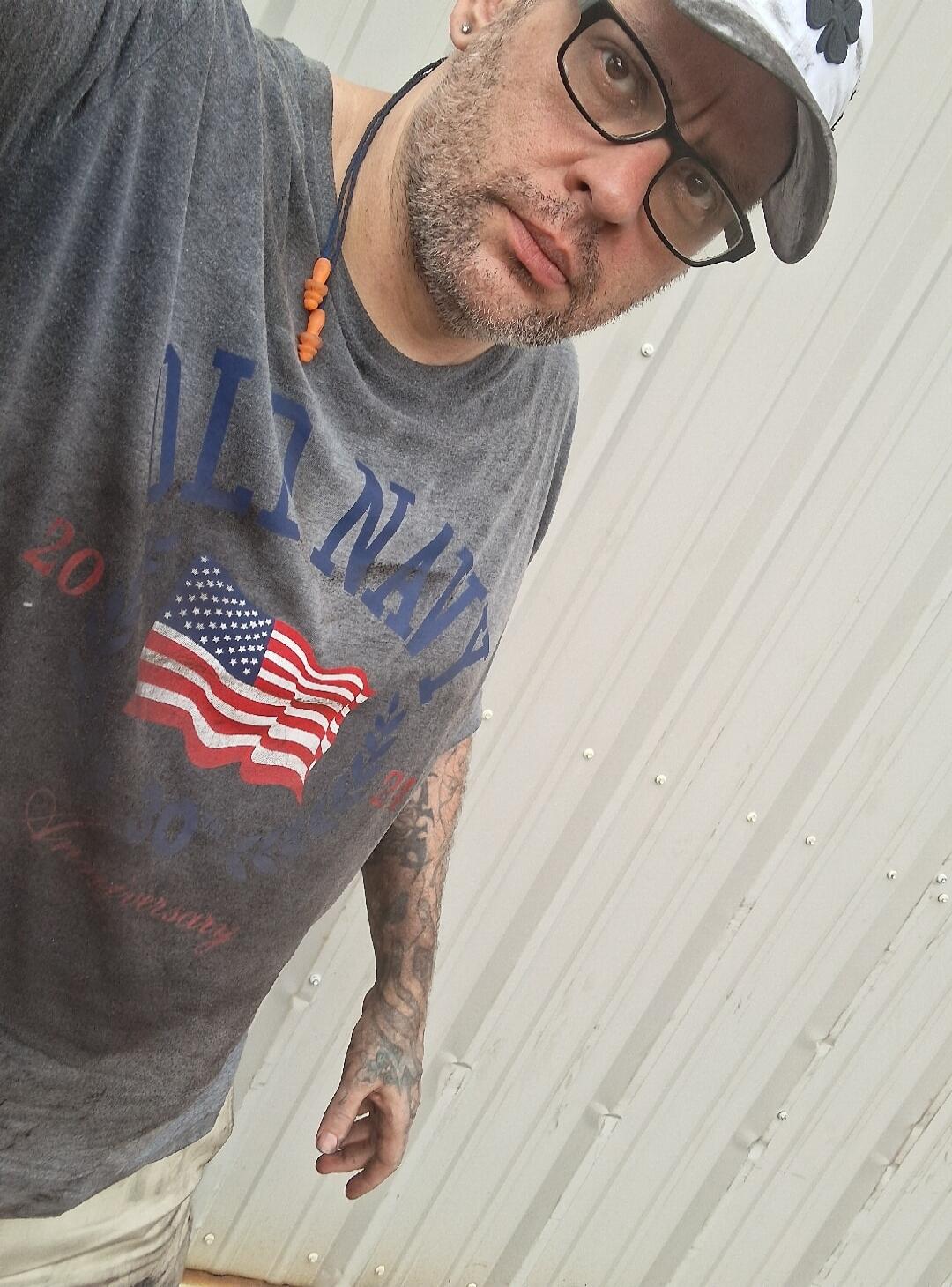 A man wearing a grey t-shirt with 'ILL NAVY' and an American flag design, along with '20' and 'Anniversary' text. He has a beard, glasses, a cap, and visible tattoos on his arm. He is looking directly at the camera.