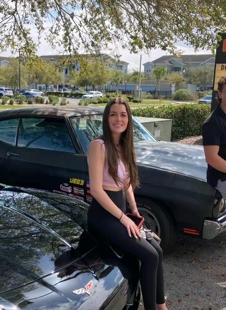 A young woman with long brown hair is sitting on the hood of a black sports car, possibly a Corvette. She is wearing a light pink crop top and black leggings. Behind her is another dark classic car with 'JEG'S' stickers. The scene is outdoors on a sunny day, with trees and buildings in the background.