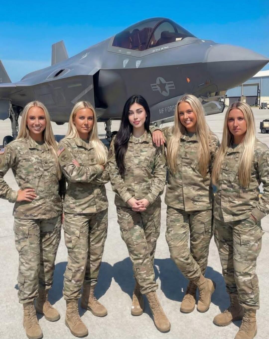 Five women in military camouflage uniforms are standing in front of a US Air Force F-35 fighter jet. The jet has 'US AIR FORCE' visible on its side. The women are posing for the camera on an airfield.