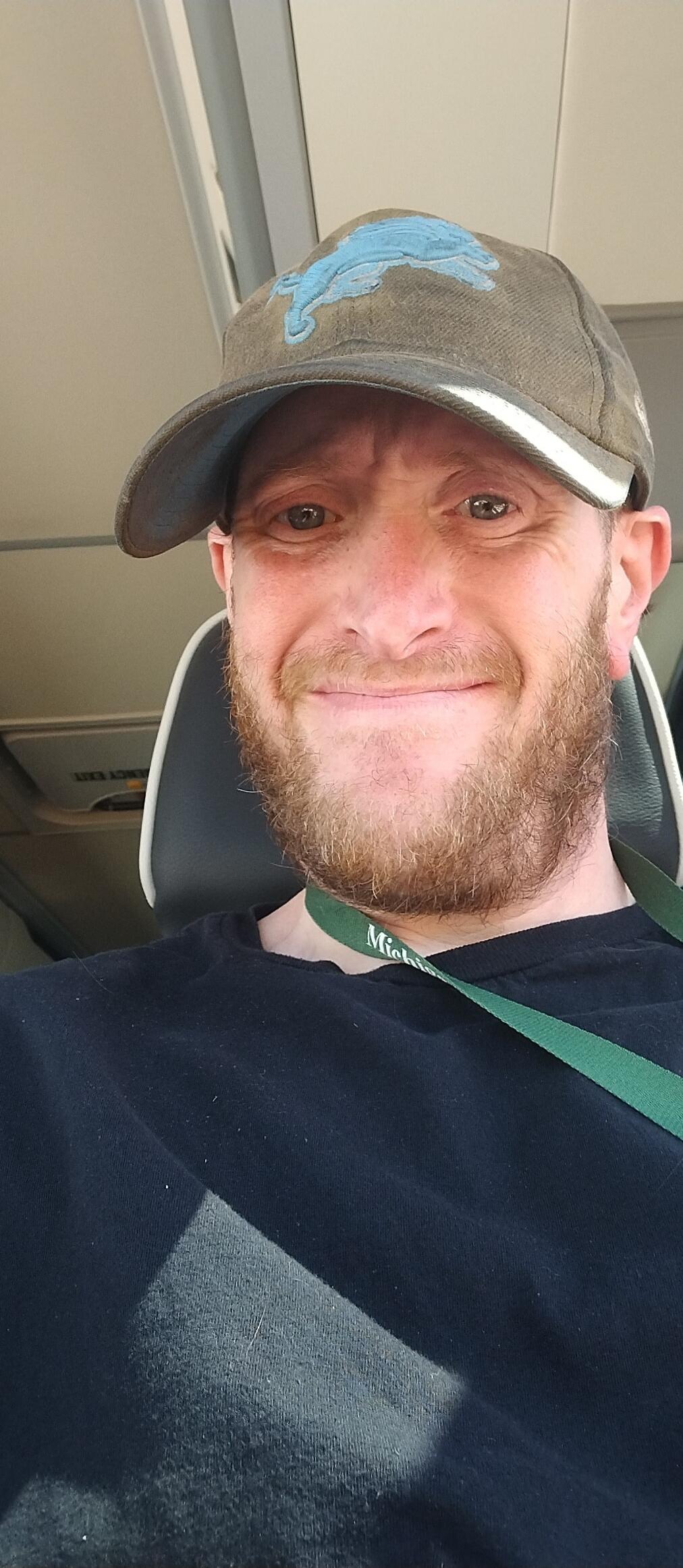 A man with a beard and a cap is smiling at the camera. He is wearing a black t-shirt and a green lanyard with 'Michig' visible. In the background, an 'EMERGENCY EXIT' sign is visible on the overhead panel. The man appears to be inside a vehicle.