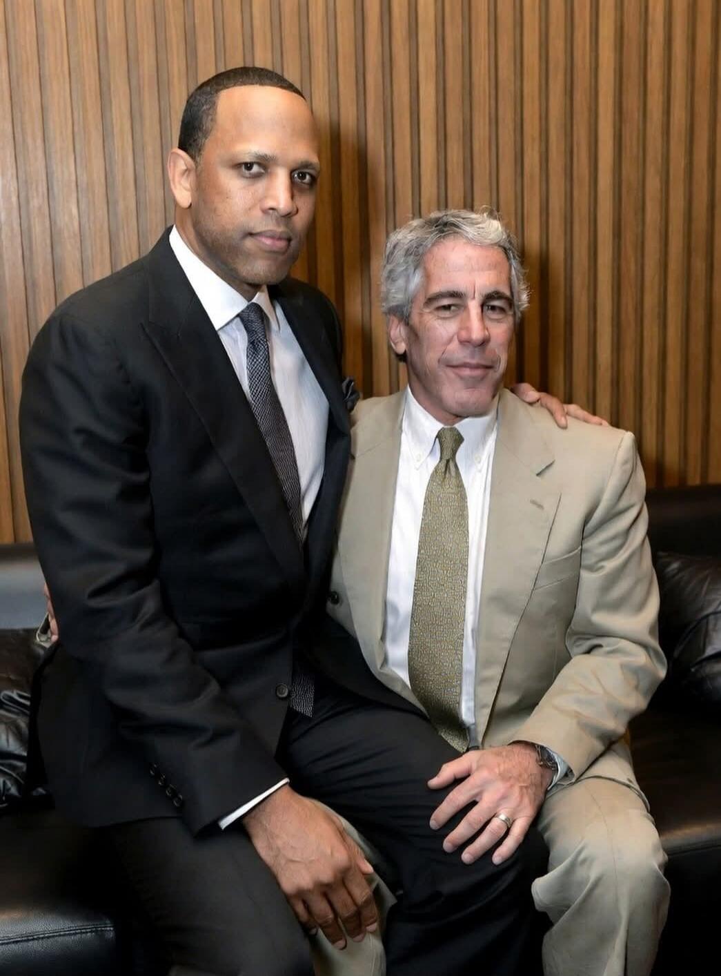 Two men in suits sitting on a black leather couch in front of a wooden paneled wall.