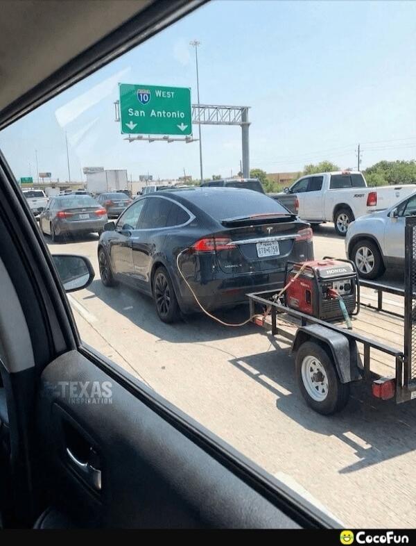 I-10 WEST San Antonio. TEXAS INSPIRATION. CocoFun. A black Tesla Model X is seen on a highway, towing a small trailer with a red Honda generator. A cable connects the generator to the Tesla, suggesting it's being used to charge the electric car.