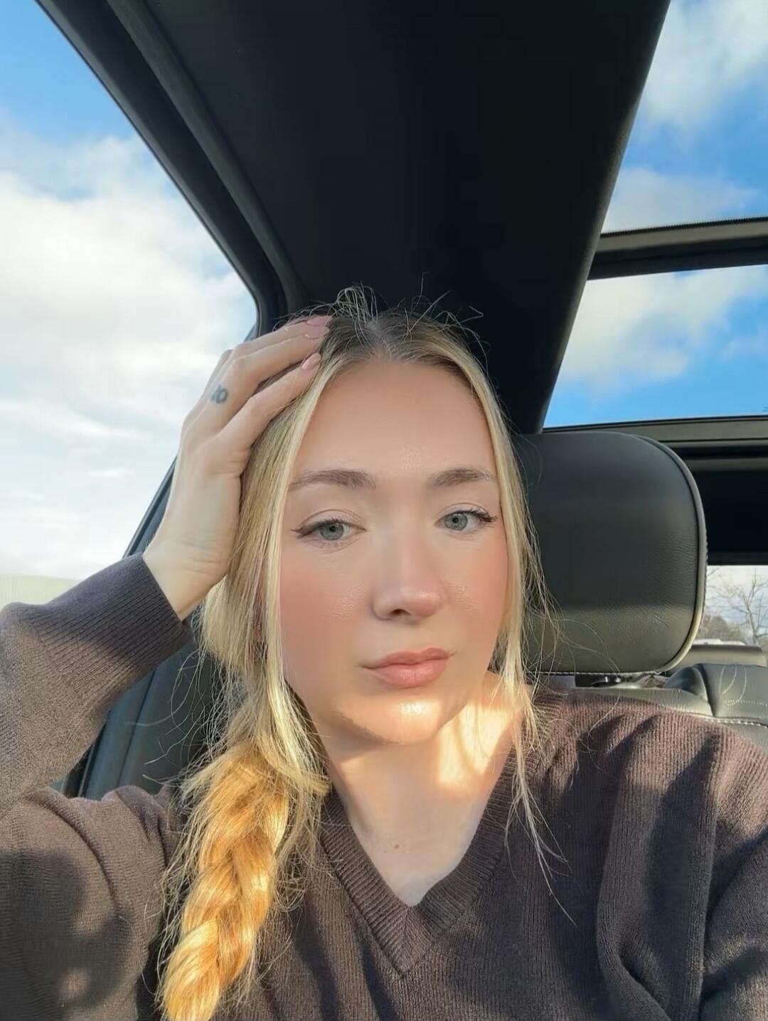 A young blonde woman with a braid, wearing a dark V-neck sweater, takes a selfie in a car. Her hand is on her head, and sunlight illuminates her face. A sunroof shows a blue sky with clouds.