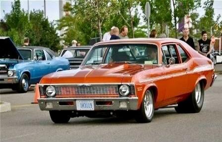 Orange classic muscle car at a car show, with people gathered in the background. A blue car is visible on the left. License plate is faintly visible on the orange car.