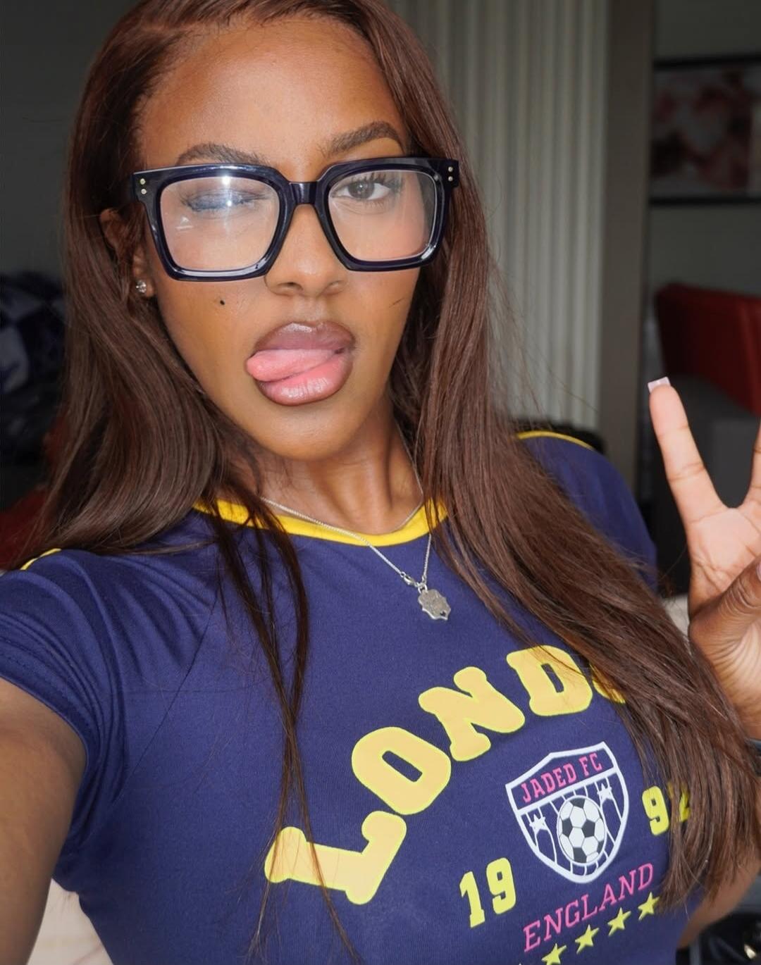A woman wearing glasses and a blue t-shirt with 'LONDON JADED FC 1994 ENGLAND' text, winking and sticking her tongue out while making a peace sign.
