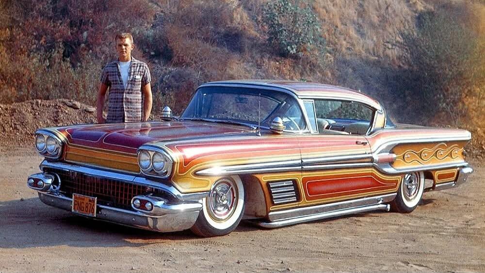 A man stands beside a customized vintage car with a metallic two-tone paint job parked on a dirt road.