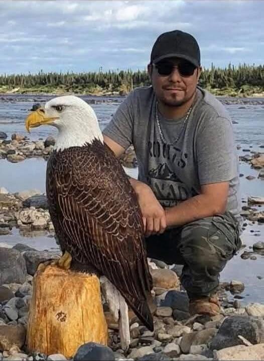 A man wearing sunglasses and a dark cap kneels beside a large bald eagle perched on a log along a rocky shore.