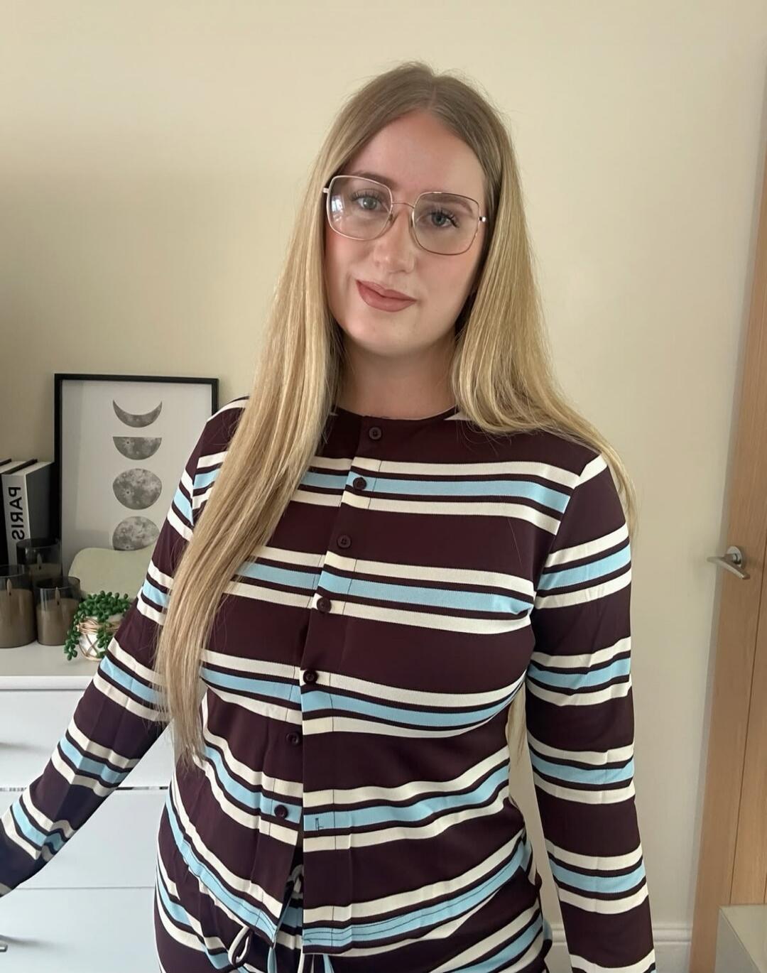 A young woman with long blonde hair and glasses is smiling at the camera. She is wearing a striped long-sleeve top with brown, light blue, and white stripes. In the background, there is a white dresser with a framed picture depicting moon phases and a book with 'PARIS' written on it.