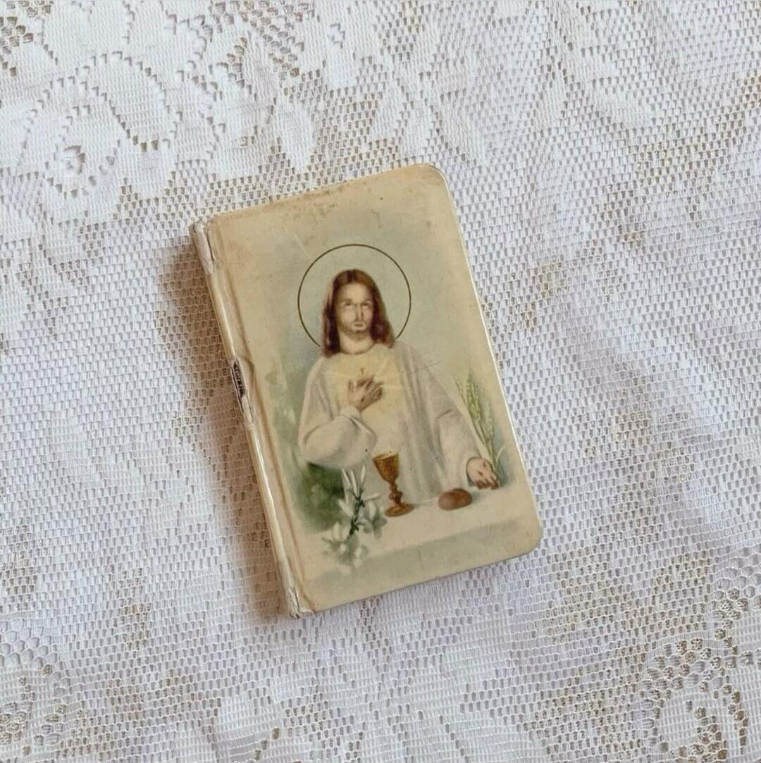 An old religious book or prayer card featuring an illustration of Jesus Christ with a halo, a glowing heart, a chalice, lilies, and bread. The item is placed on a white lace tablecloth.