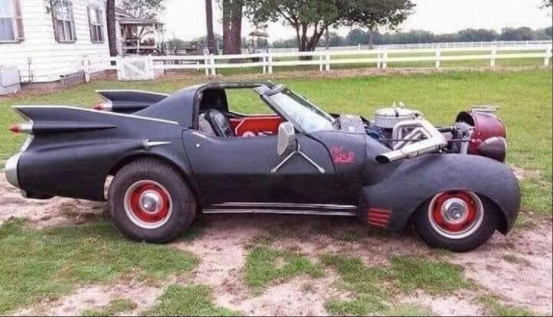 A highly customized black car with large tail fins and an exposed engine, resembling a hot rod or a modified Batmobile, parked on a grassy area next to a house.