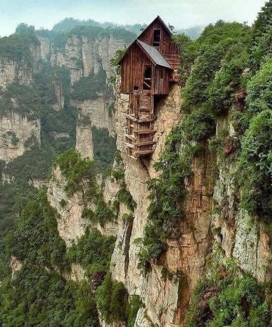 A wooden hut perched on the edge of a cliff, built on a narrow rock pillar with surrounding trees and a dramatic drop.