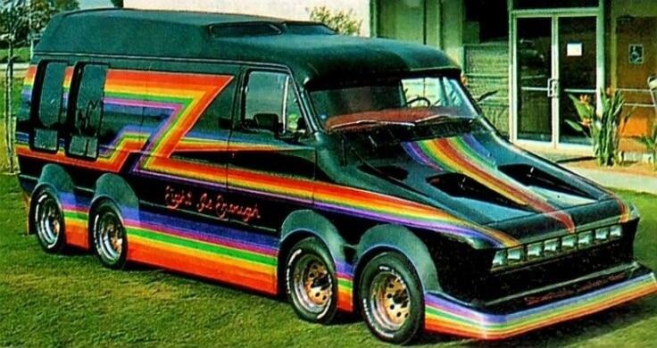 A highly customized, elongated black van with a vibrant rainbow stripe paint job, featuring multiple wheels on each side. The text 'High Roller' is visible on the side. The van is parked on a grassy area in front of a building.