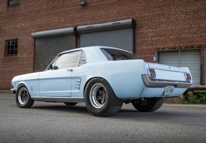 A light blue classic Ford Mustang with wide rear tires, parked in front of a brick building with garage doors. The license plate reads 104 88H.
