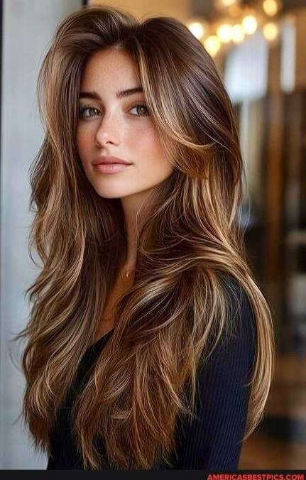 A woman with long, wavy brown hair with caramel highlights, wearing a dark top and looking slightly to the side.