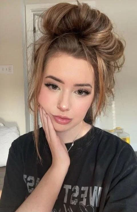 A young woman with a messy bun, wearing a dark t-shirt with the word 'WEST' (mirrored) on it, posing with her hand on her cheek. She has makeup and a nose piercing.
