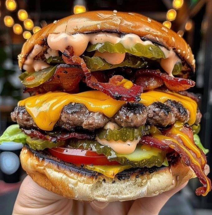 A towering, overloaded cheeseburger with multiple patties, melted cheese, bacon, pickles, onions, and various sauces stacked between a glossy bun.