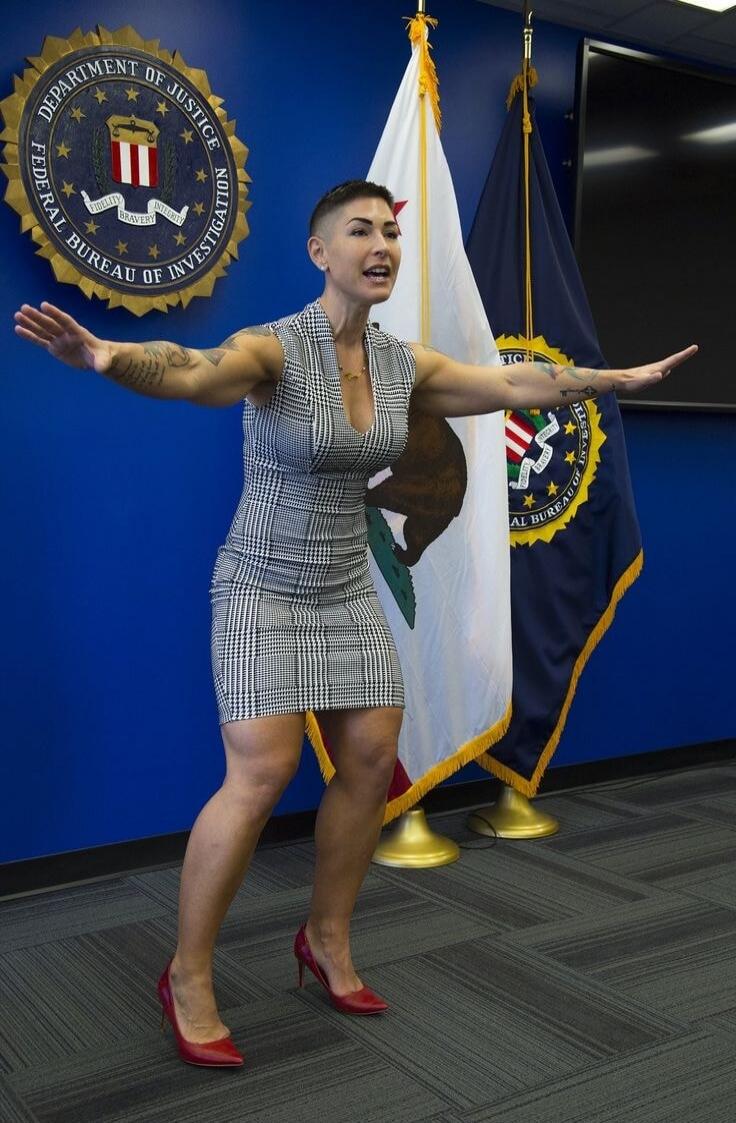 DEPARTMENT OF JUSTICE FEDERAL BUREAU OF INVESTIGATION FIDELITY BRAVERY INTEGRITY. A woman with a distinctive haircut and tattoos is speaking or presenting in front of official seals and flags.