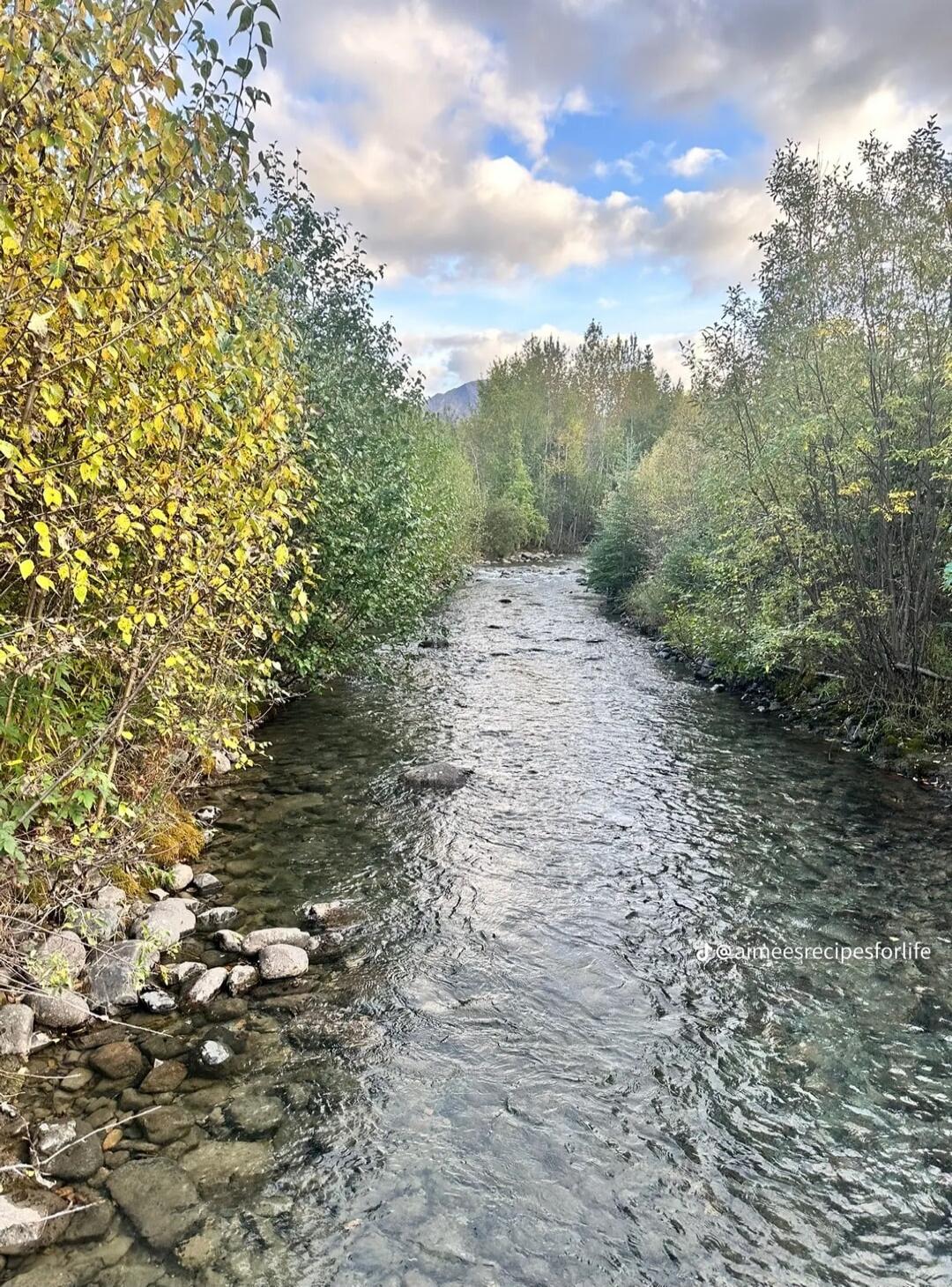 A tranquil river flowing between rocky banks, lined with trees showing autumn colors. The water is clear, and the sky above is partly cloudy with patches of blue.