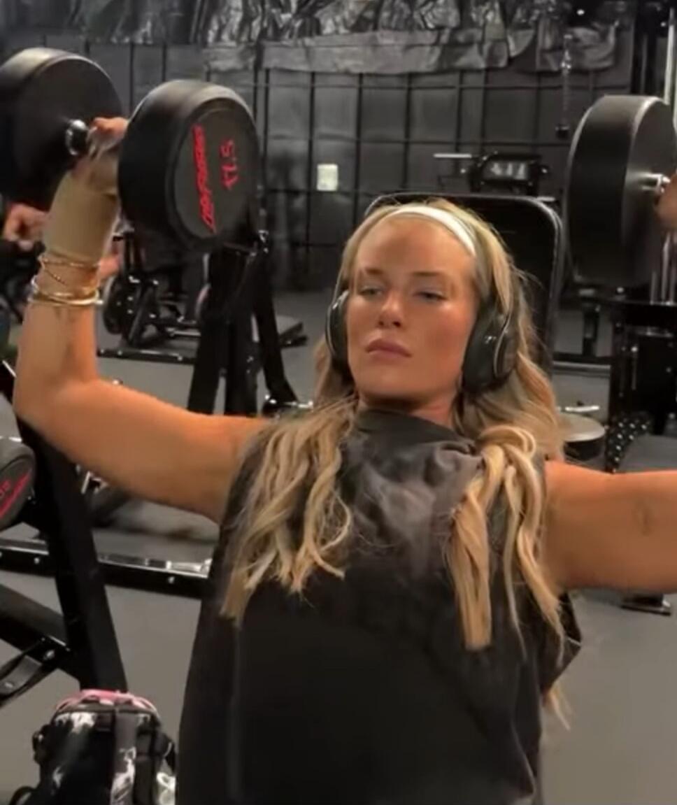 A woman in a gym, wearing headphones and a headband, is lifting dumbbells. The dumbbell has 'LifeFitness 17.5' written on it.