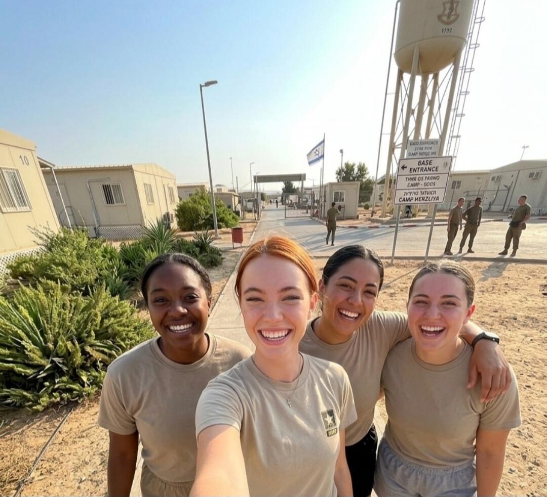 Four smiling women in military-style t-shirts take a selfie at what appears to be a military base. Signs in the background indicate 'BASE ENTRANCE' and 'CAMP HERZLIYA'. An Israeli flag is visible. One woman's shirt has a 'U.S. ARMY' logo. A water tower with '1777' is also visible.