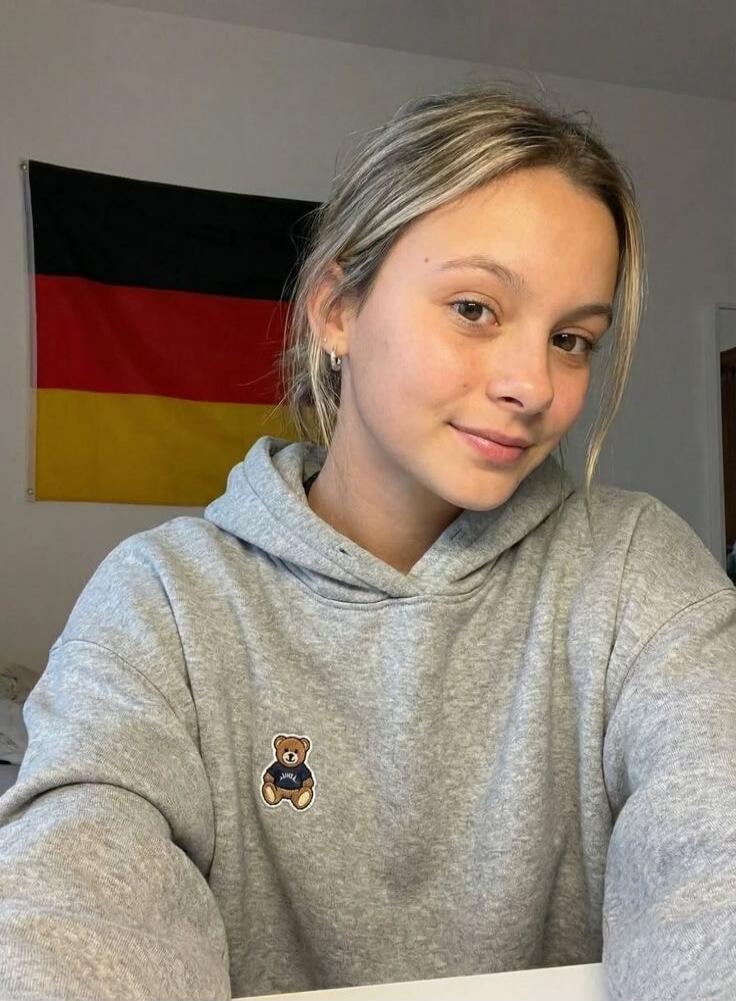 A young woman with blonde hair, wearing a grey hoodie with a teddy bear patch, smiles at the camera. A German flag is visible in the background.