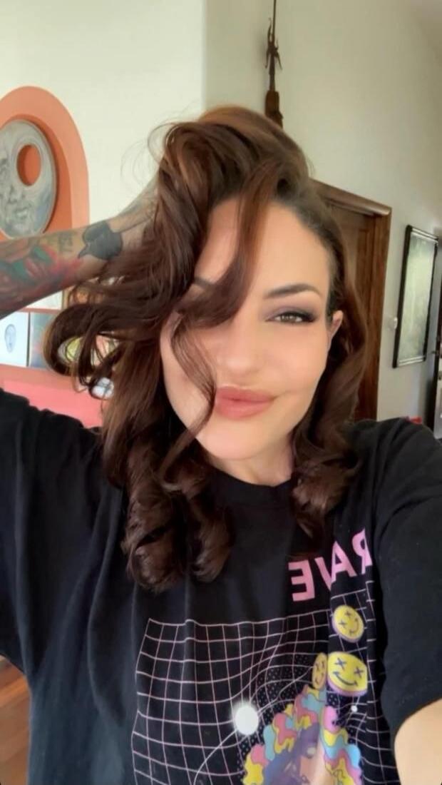A woman with dark, curly hair and tattoos on her arm, smiling at the camera. She is wearing a black t-shirt with a grid pattern and colorful graphics, including smiley faces. Mirrored text is visible on the shirt.